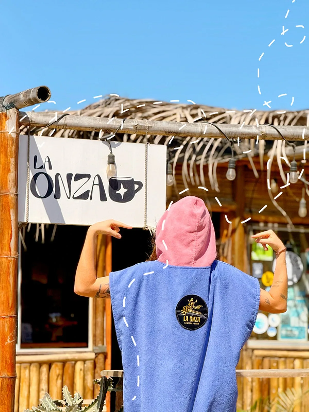 Una persona con un poncho azul y capucha rosada con el parche de La Onza Café, frente al letrero del café en Lobitos